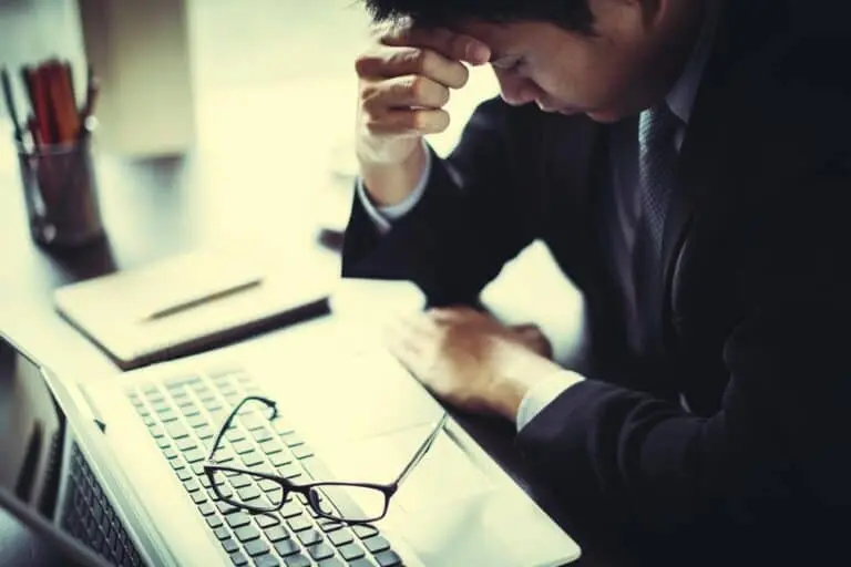 A man with his head in his hand with his eyes close, looking very frustrated. His glasses are on top of his laptop.