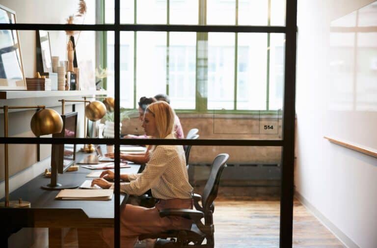 Three employees working at their computers at a communal desk. The view is through a window wall.