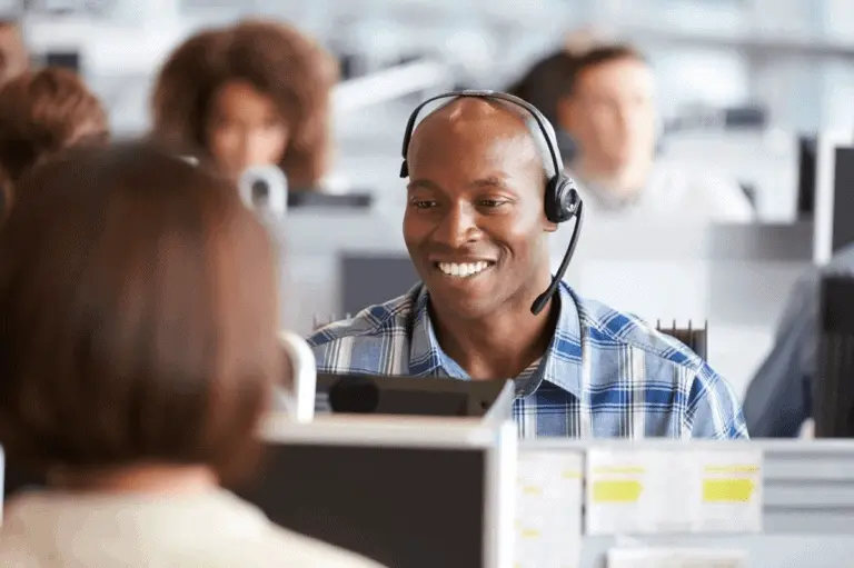 Man working on his computer in a cubical style office with a headset on. Other employees working around him out of focus.