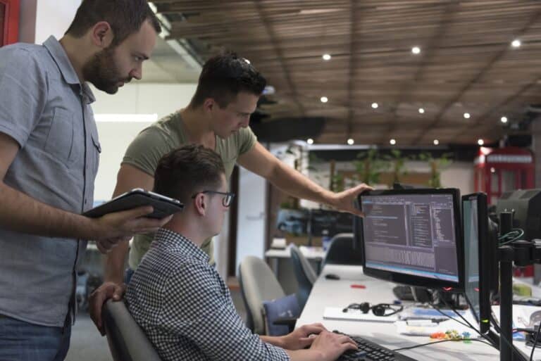 3 young employees looking at information on a computer. One is sitting down typing on the computer, the other two are standing behind him, one of them is holding a tablet.