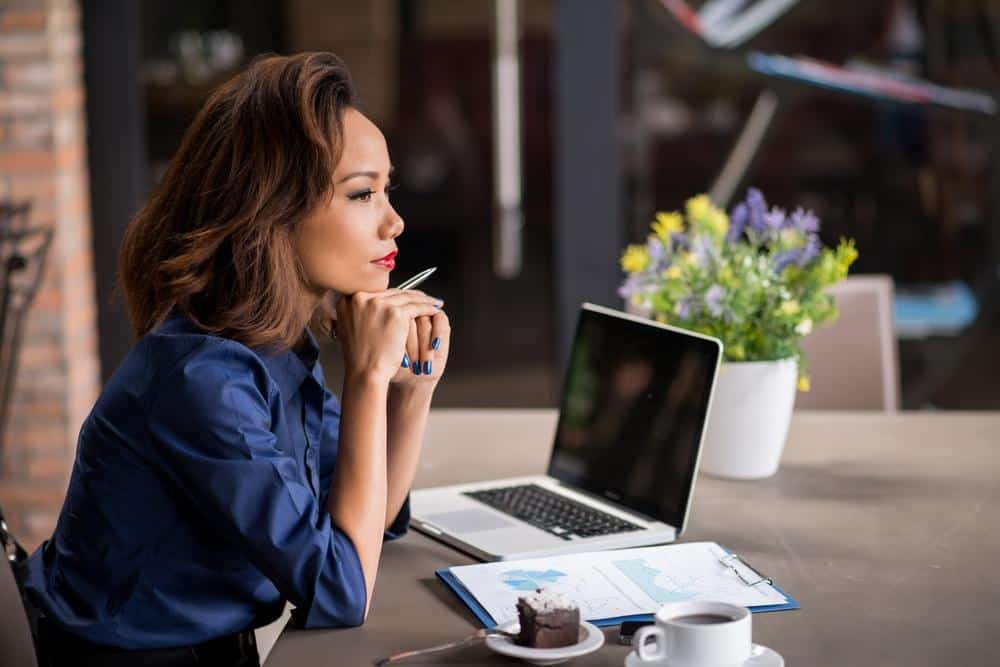 Woman sitting at a table with her laptop and a clipboard , staring off into the distance thinking. There is a cup of coffee and piece of cake on the table as well.