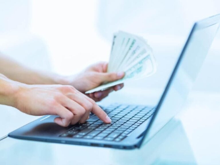 Close up of a person typing on a laptop with a handful of one hundred dollar bills in her other hand.